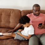 A man sitting on a sofa with this son. They are both using smartphones (Photo: August de Richelieu)